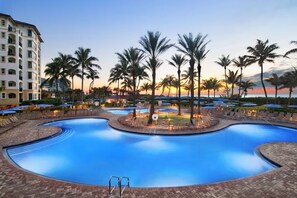Una grande piscina a forma di curva circondata da palme e lettini, con un edificio sullo sfondo.