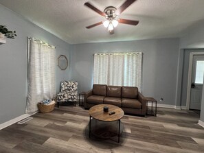 Un salon avec un canapĂ© en cuir marron, une table basse ronde, un ventilateur de plafond et une fenĂȘtre avec des rideaux.