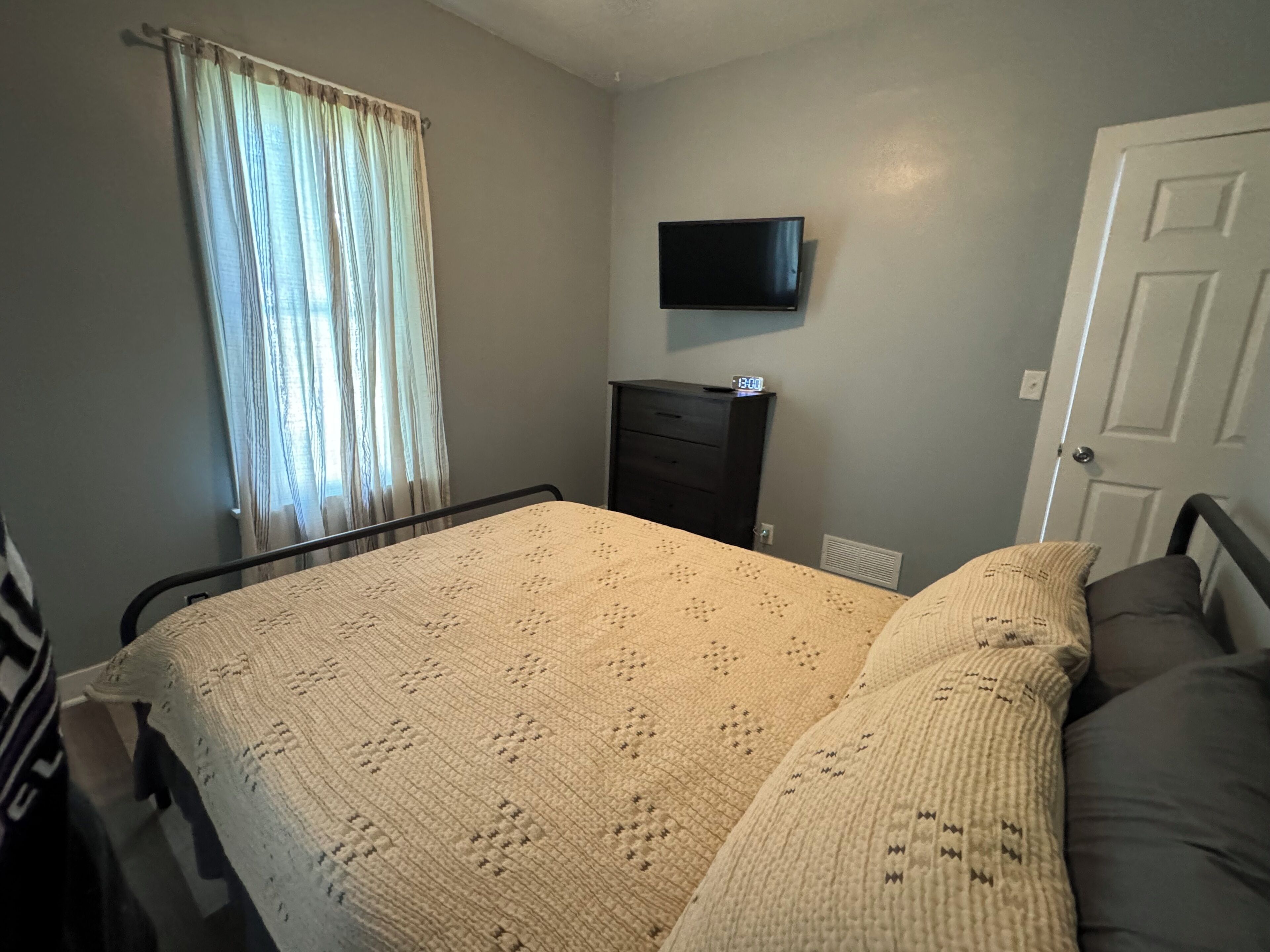 Une chambre à coucher avec un lit, un téléviseur fixé au mur, une commode et une fenêtre avec des rideaux.