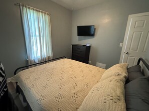Une chambre Ă coucher avec un lit, un tĂ©lĂ©viseur fixĂ© au mur, une commode et une fenĂȘtre avec des rideaux.