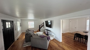 A spacious living room with a grey sofa, a round ottoman, and a TV mounted on the wall. - Newly remodeled Spacious 4-BM house in Phoenixville with OS parking/ central, AC (Phoenixville)
