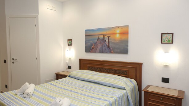 A hotel room with a bed, a nightstand, a wooden dresser, and a painting of a sunset over a pier.