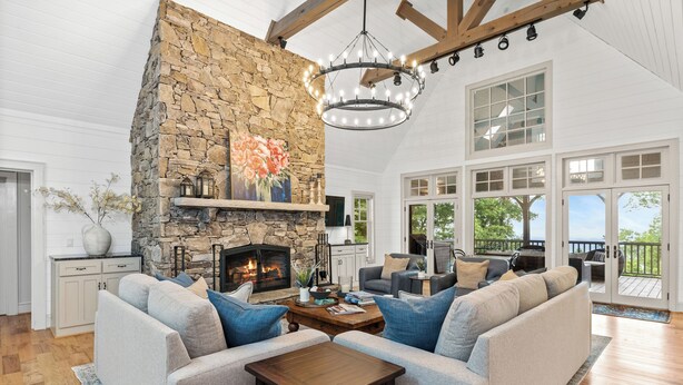 A spacious living room with a stone fireplace, a chandelier, and a large window overlooking a deck. - Luxury Home. Blue Valley Views. Extraordinary Attention to Detail | Sevenoaks Cottage (Highlands)