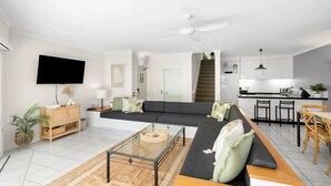 A modern living room with a sofa, coffee table, and TV. There is a staircase leading to a kitchen with bar stools.