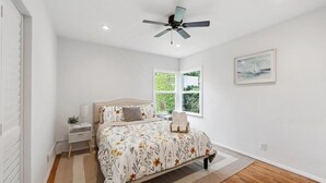 Un dormitorio con una cama, un ventilador de techo, una ventana y un cuadro en la pared.