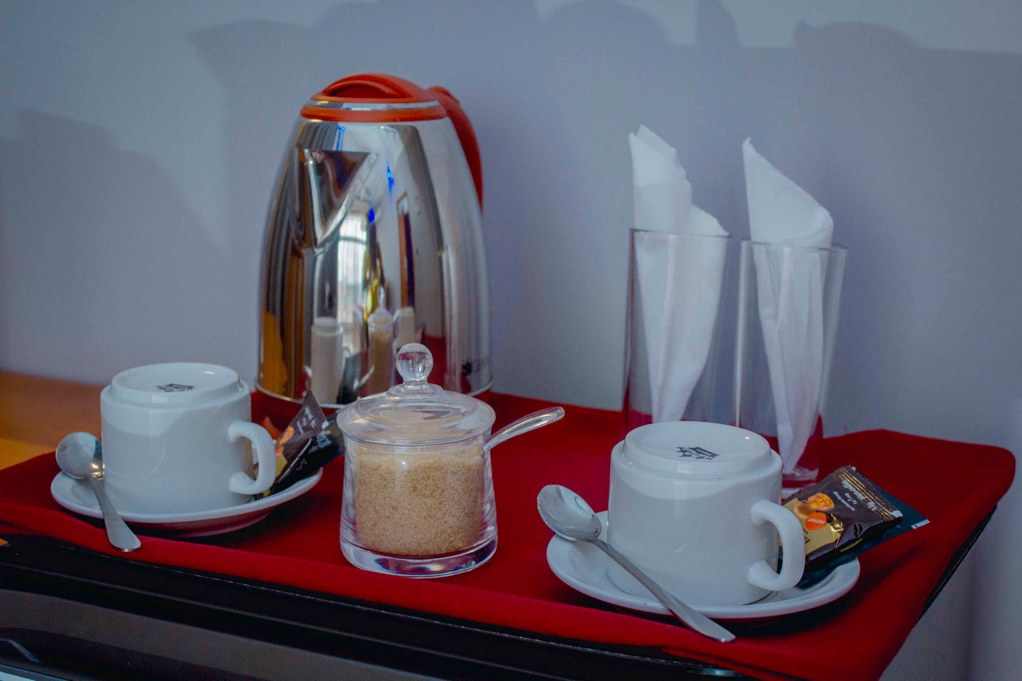 Disposição para o café da manhã com duas xícaras brancas, um pote de vidro com açúcar, uma cafeteira e um sachê de creme em um tabuleiro vermelho.