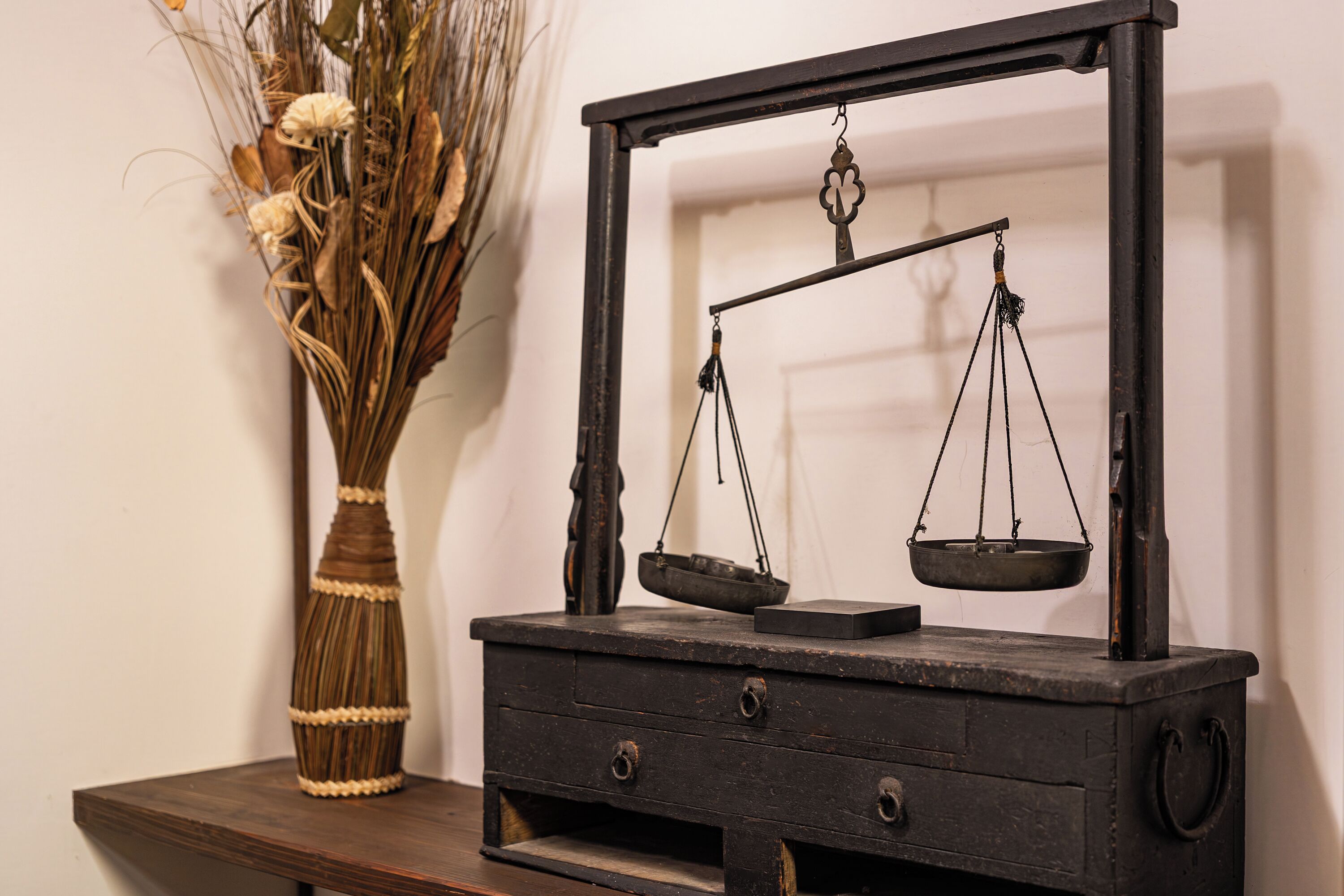 Una balanza antigua sobre una mesa de madera, con un jarrón de flores secas a su lado.