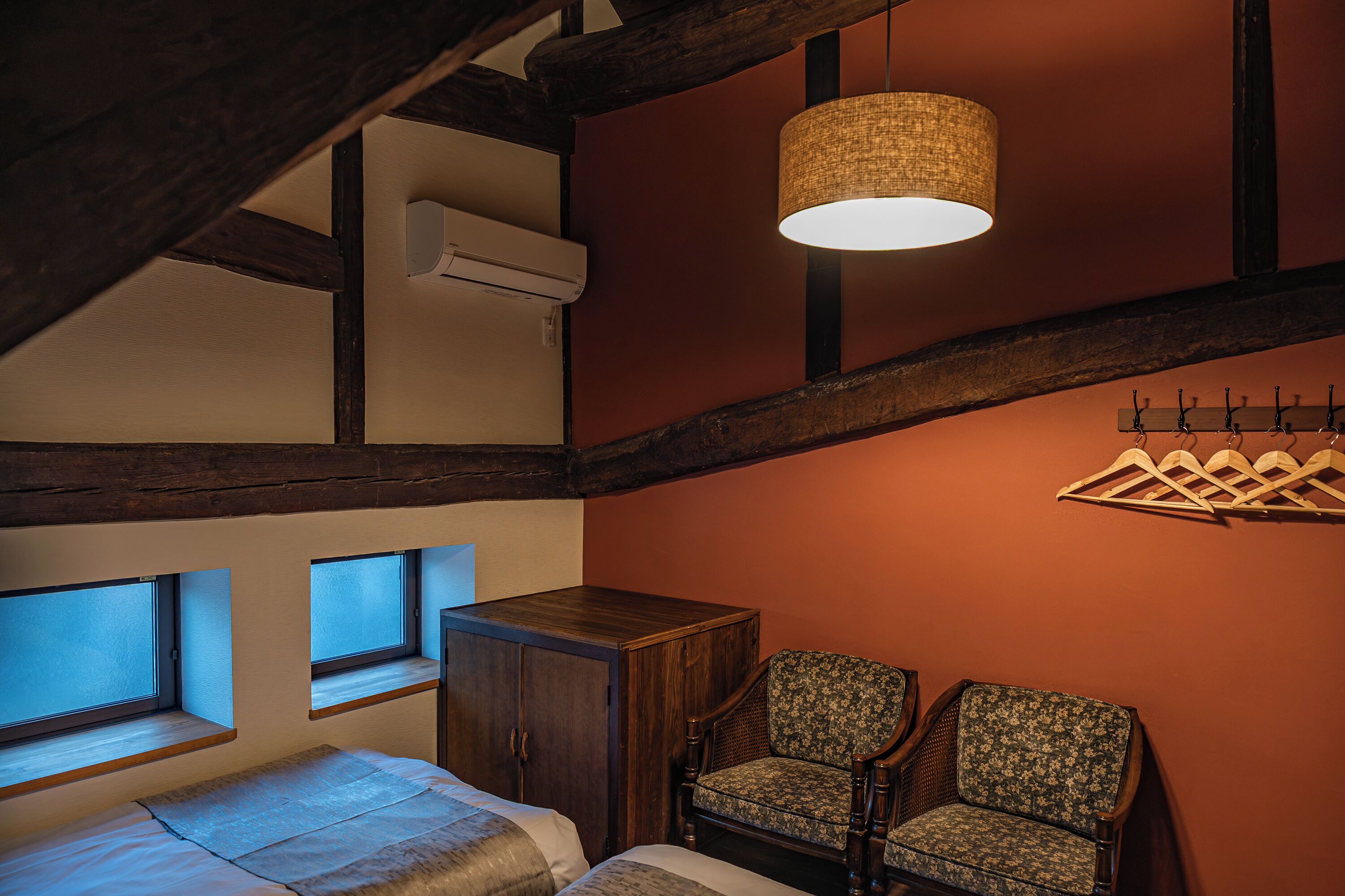 A cozy room with a bed, two chairs, a wooden cabinet, a hanging light fixture, and a coat rack.