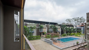 A modern outdoor pool area with a wooden deck, a covered seating area, and a view of a residential building in the background. - Alaya Stays 3-BHK with Pool near Khandala Hills - Imperial Serenia (Lonavala)