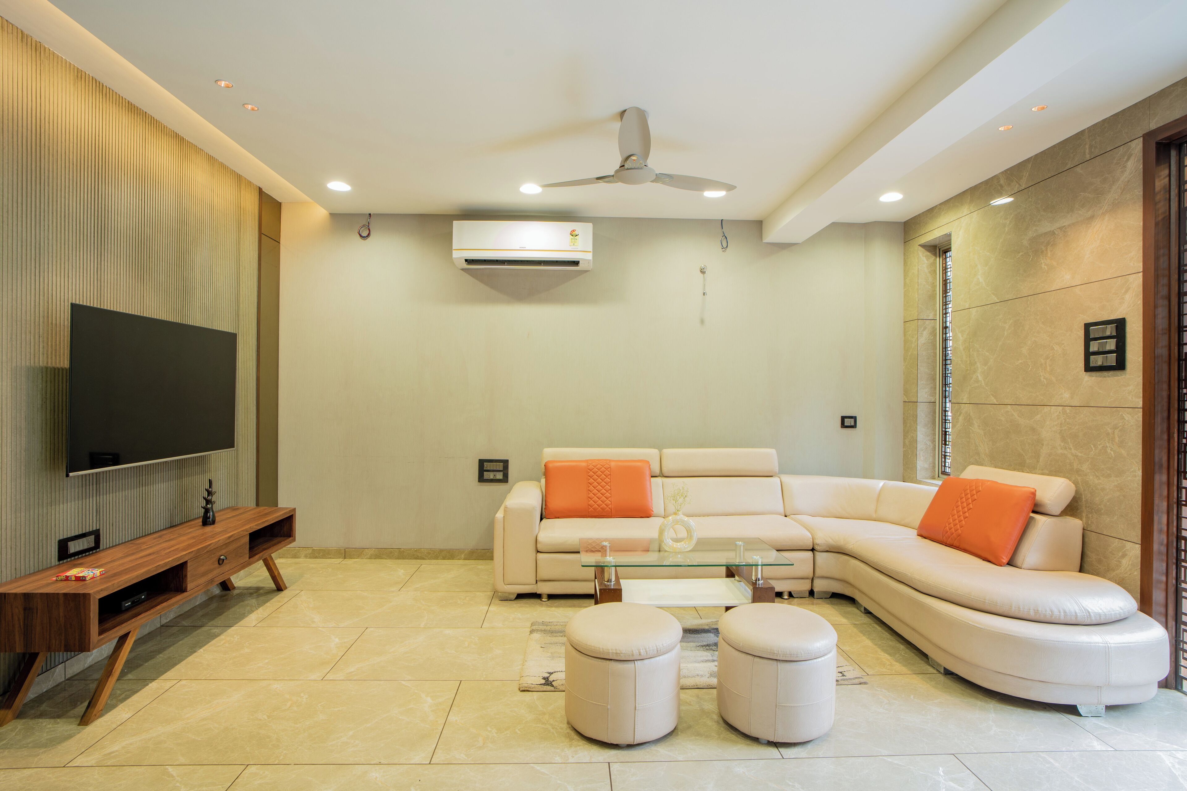 Una sala de estar moderna con un televisor de pantalla plana, un sofá modular con cojines naranjas, una mesa de centro de vidrio y un ventilador de techo.