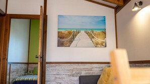 Quarto com teto de madeira, pintura de um caminho de praia, cama com travesseiro verde e sofá cinza com manta amarela.