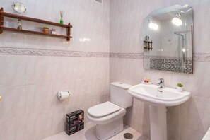 Une salle de bain avec des toilettes, un lavabo et un miroir. On y trouve une étagère avec des articles de toilette et une boîte contenant un sèche-cheveux, posée sur le sol.