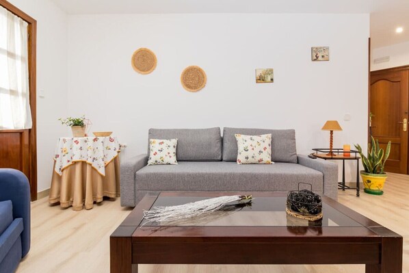 Un salon avec un canapé gris, des coussins à motifs floraux, une table basse agrémentée d’objets décoratifs, une petite table d’appoint avec une lampe et une plante en pot.