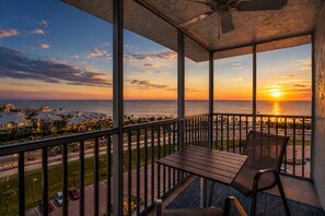 A balcony with a view of the sunset over the ocean, a table, and chairs.