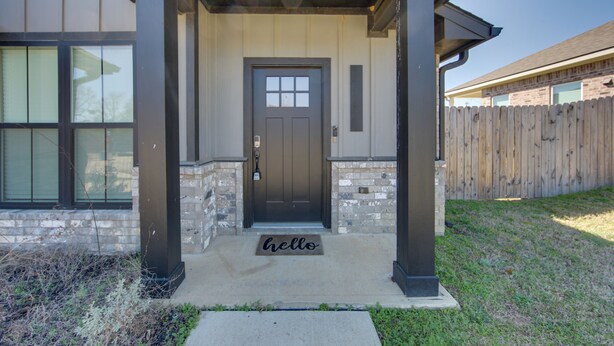 Une entrée de maison avec une porte marron foncé, un paillasson où est écrit « Bonjour », et une façade en pierre et en briques.
