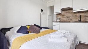 A modern bedroom with a neatly made bed, a bedside table, and a kitchen area with white cabinets and a brick backsplash.