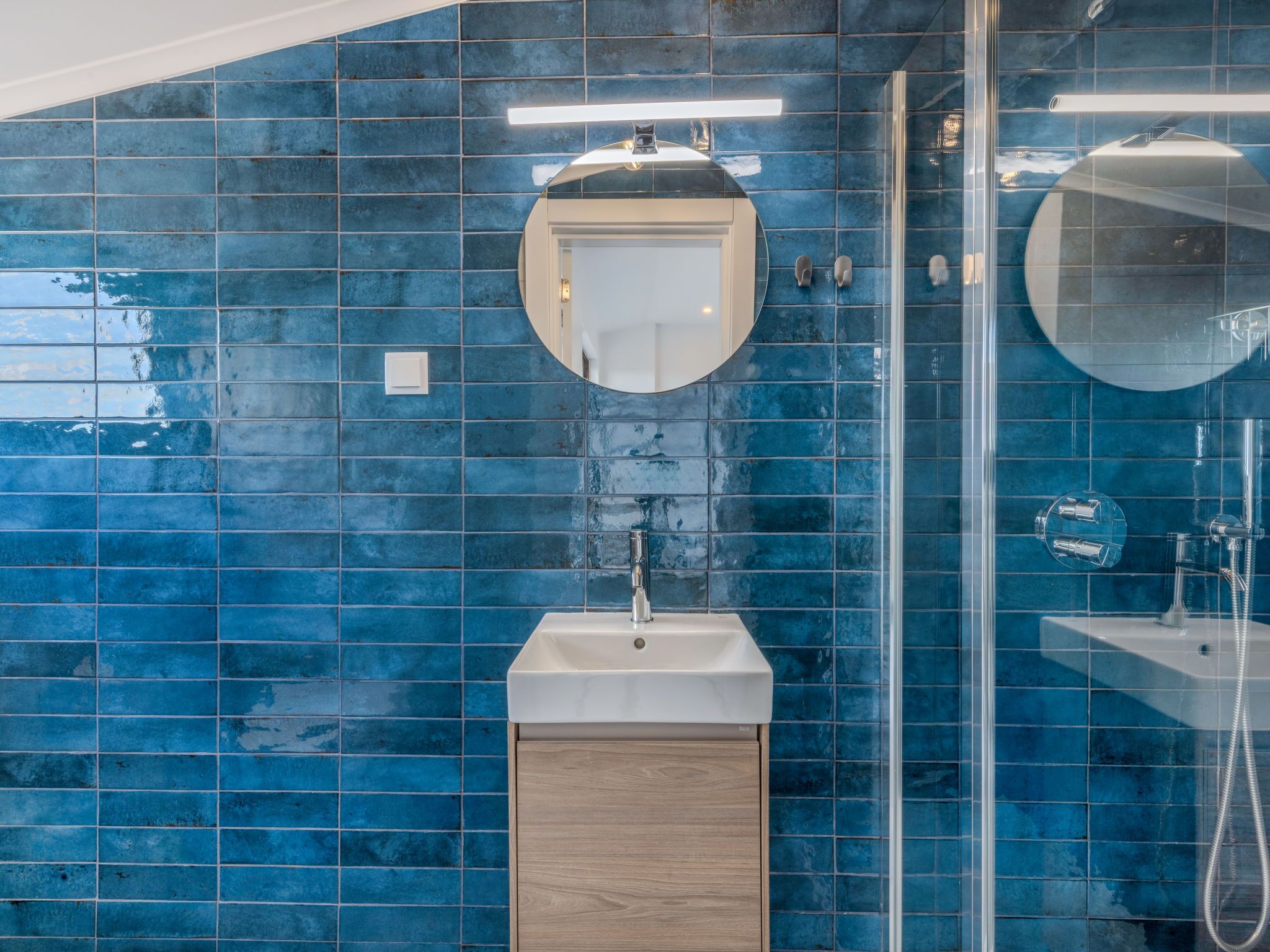A bathroom with blue tiles, a round mirror, a sink, and a shower area.