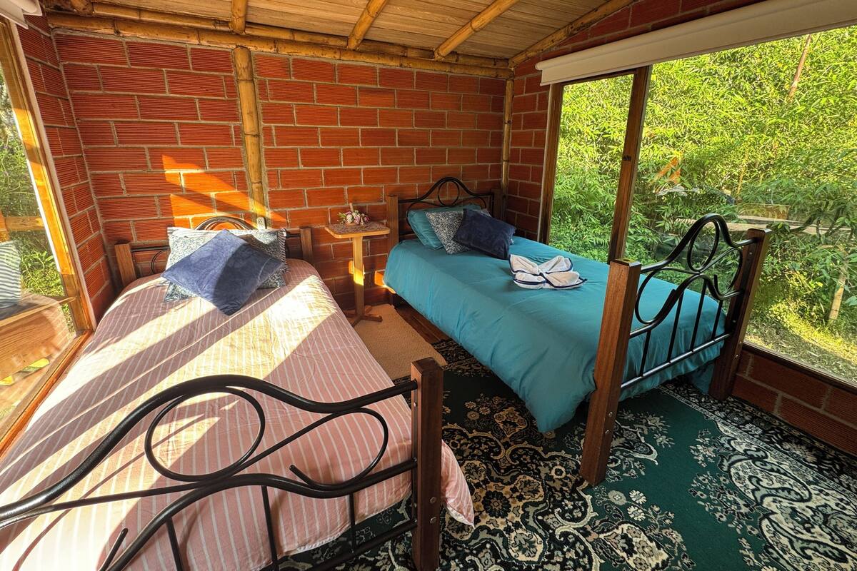 A room with two beds, a wooden ceiling, brick walls, and a view of greenery outside.