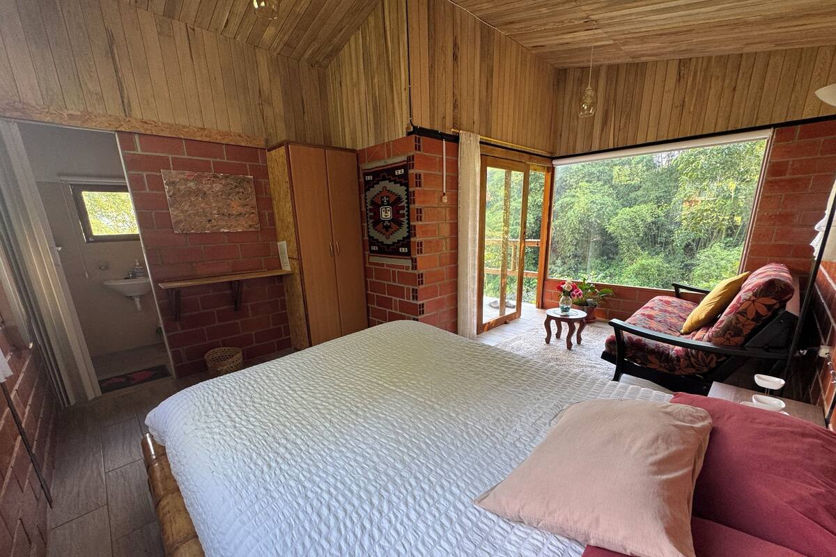 A bedroom with a large bed, a wooden ceiling, a brick wall, a window, and a balcony with a chair and table.