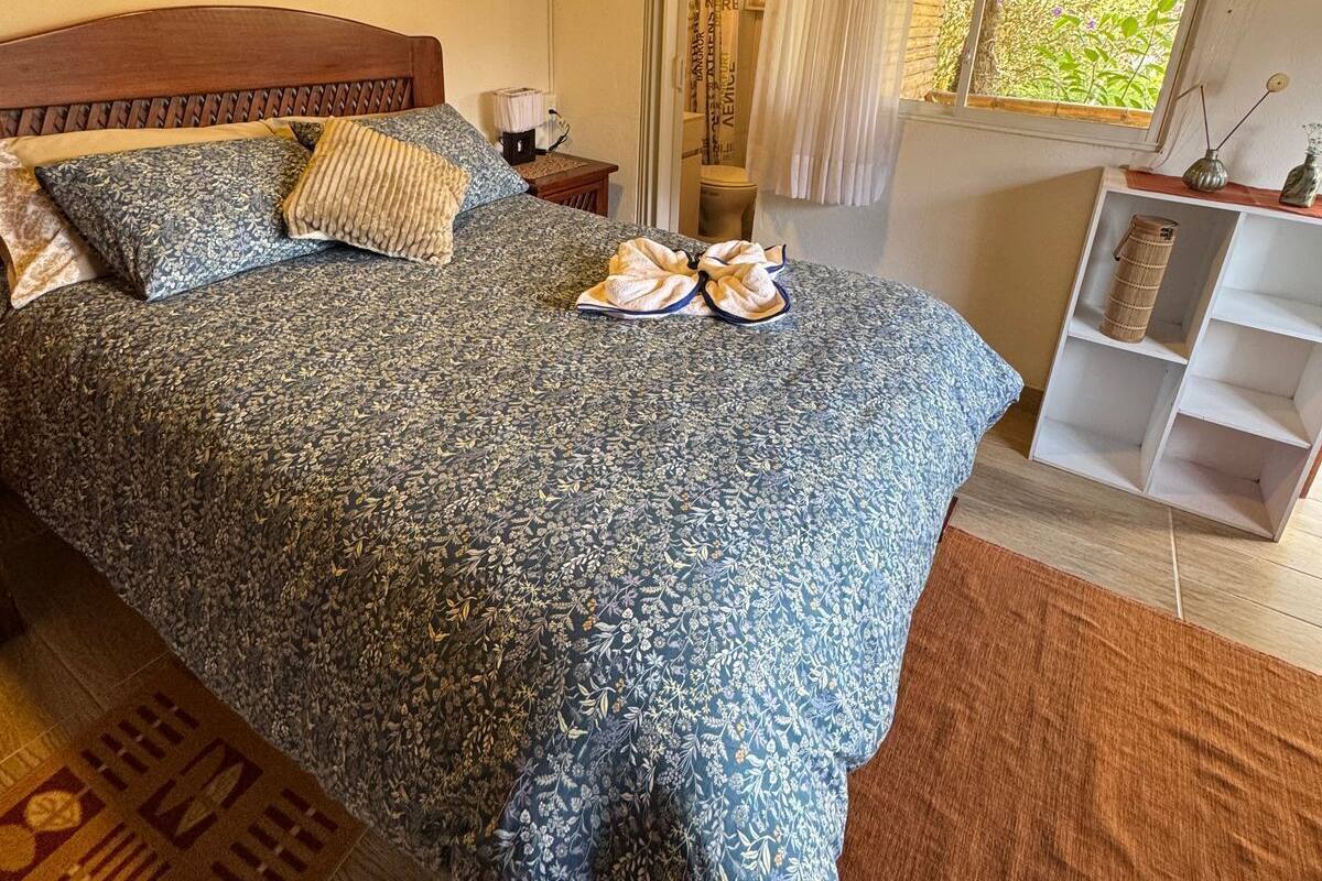 A bedroom with a wooden ceiling, a bed with patterned bedding, a nightstand, a window with curtains, and a wall-mounted artwork.