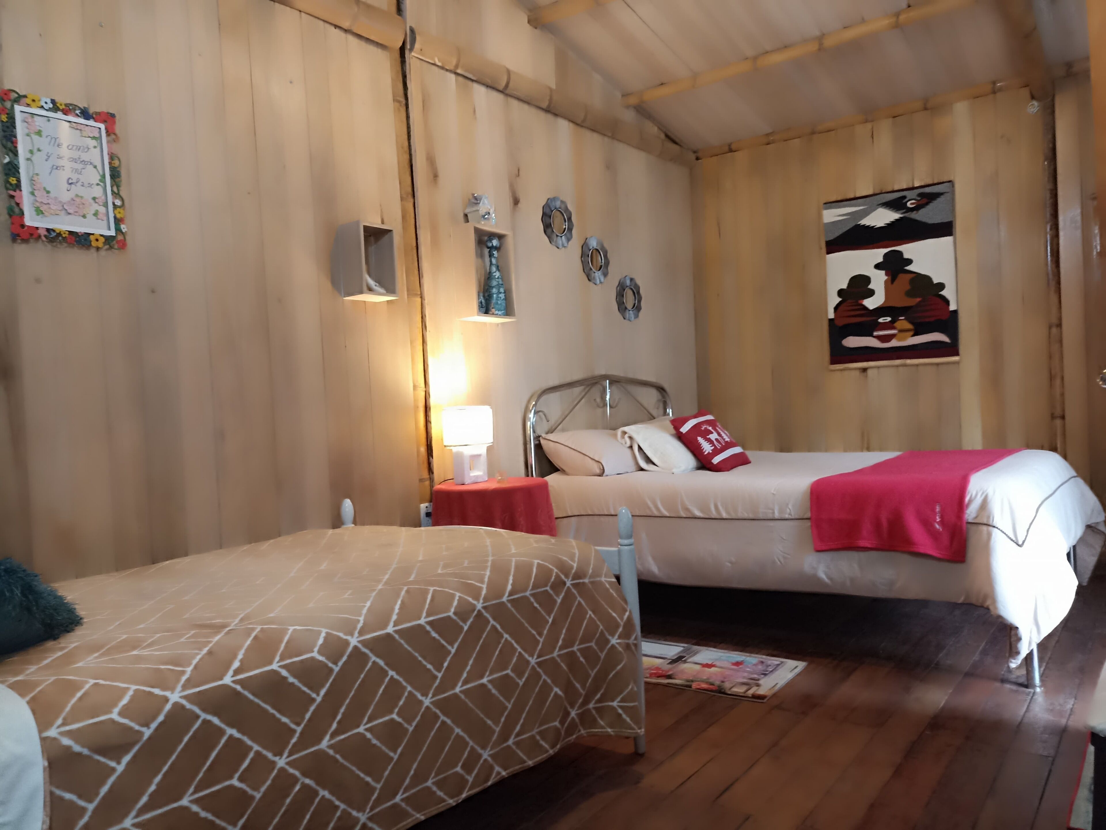 Quarto com duas camas, paredes de madeira, uma mesinha com abajur e objetos decorativos.