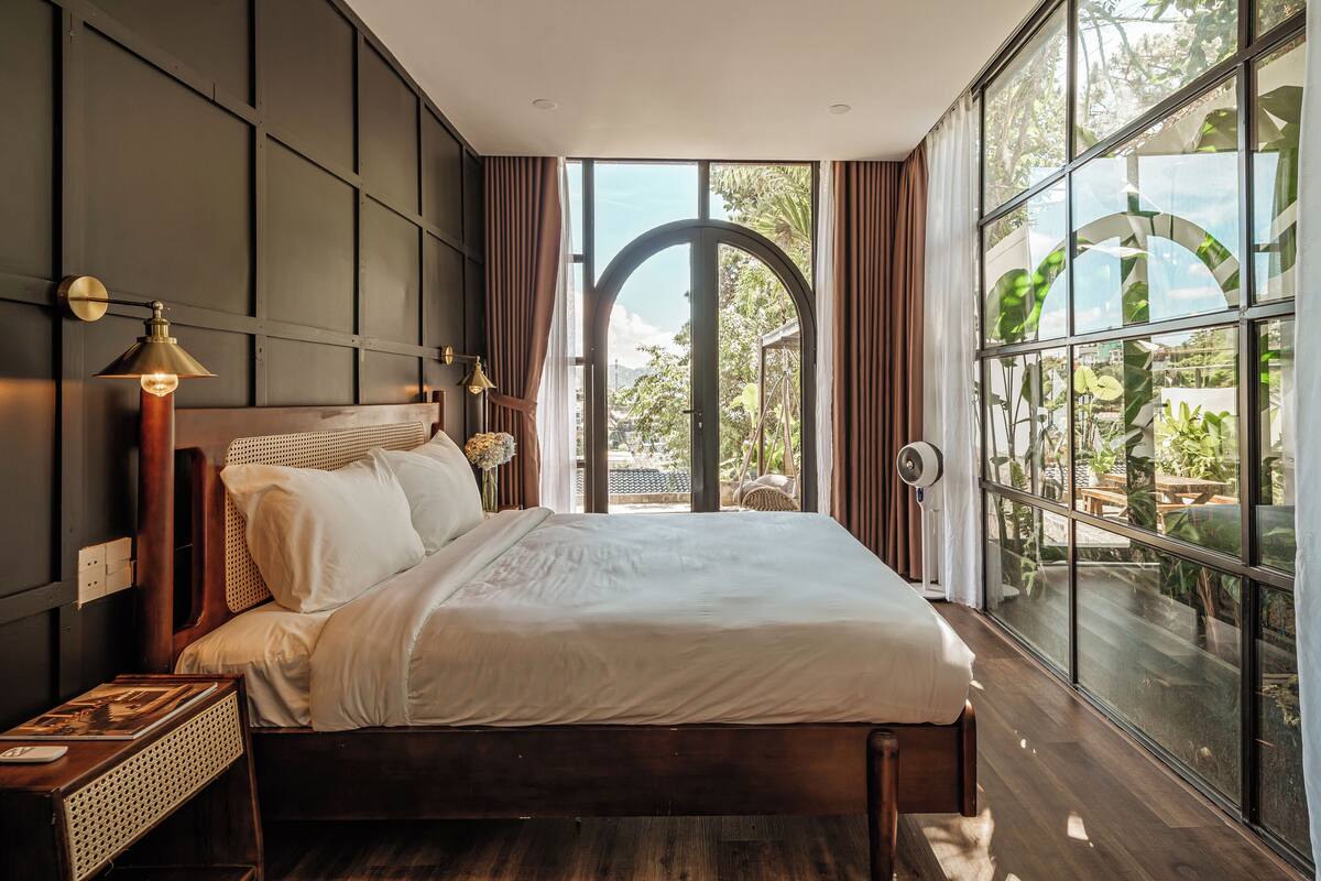 Een moderne slaapkamer met een groot bed, een houten hoofdeinde en uitzicht op groen via grote ramen.