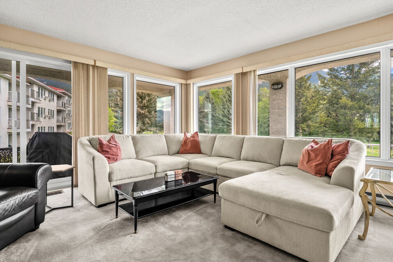 A modern living room with a sectional sofa, a coffee table, and large windows offering a view of a building and trees.