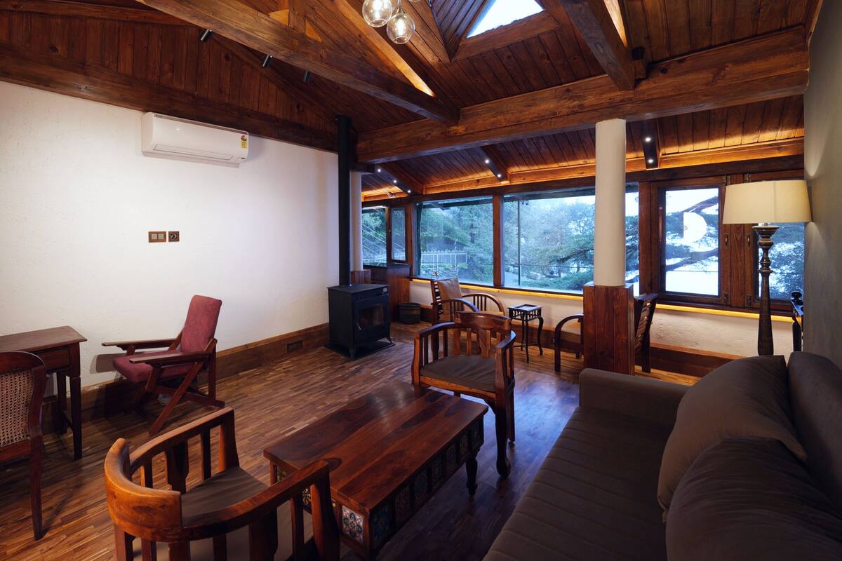 A cozy room with wooden floors, a sofa, a table, chairs, a fireplace, and large windows offering a view of trees.