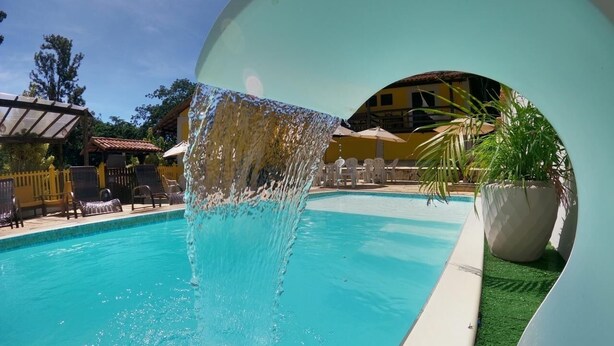 A swimming pool with a water feature, surrounded by lounge chairs and umbrellas. - Pousada alto das arvores (Buzios)