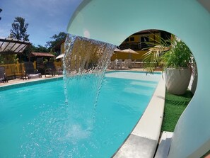 Ein Schwimmbecken mit Wasserspiel, umgeben von Liegestühlen und Sonnenschirmen.