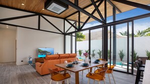A spacious living area with a wooden dining table, orange sofa, and large windows offering a view of a swimming pool and greenery. - New 19m Eco Pool Villa Sunset Garden 10 (Rawai)