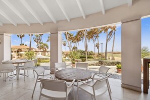 Une terrasse extérieure avec une table ronde et des chaises blanches, entourée de palmiers et offrant une vue sur un bâtiment doté d’un toit en tuiles.