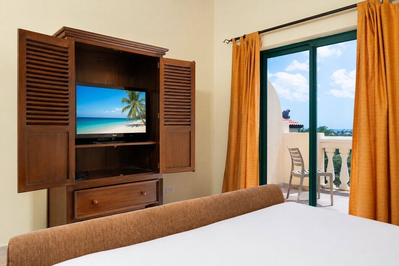Ein Hotelzimmer mit Fernseher, der eine Strandansicht zeigt, einem Balkon mit einem Stuhl und Vorhängen.