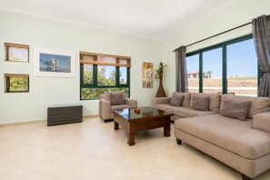 A living room with a beige sofa, a wooden coffee table, and a chair. There are two windows with blinds, a radiator, and a vase with flowers. - 3BR Villa • Steps from Pool, Restaurant &Clubhouse (Noord)