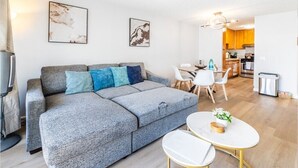 Ein modernes Wohnzimmer mit einem grauen Sofa, einem kleinen runden Tisch und weißen Stühlen. Im Hintergrund ist eine Küchenzeile zu sehen.
