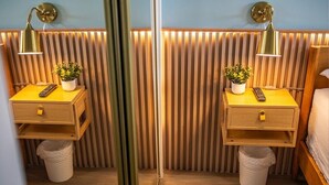 Ein Zimmer mit Spiegel, zwei Nachttischen, jeweils mit einer Lampe, einer kleinen Pflanze und einer Fernbedienung. An den Wänden sind Holzleisten angebracht und der Boden ist aus Holz.