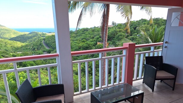 A balcony with wicker furniture, a glass table, and a view of lush greenery and distant hills. - Beautiful modern retreat with stunning views of mountain ranges & Seaview (Plymouth)