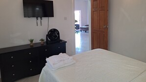 A bedroom with a bed, a TV mounted on the wall, a wooden door, and a dresser with a fan and a small plant. - Beautiful modern retreat with stunning views of mountain ranges & Seaview (Plymouth)