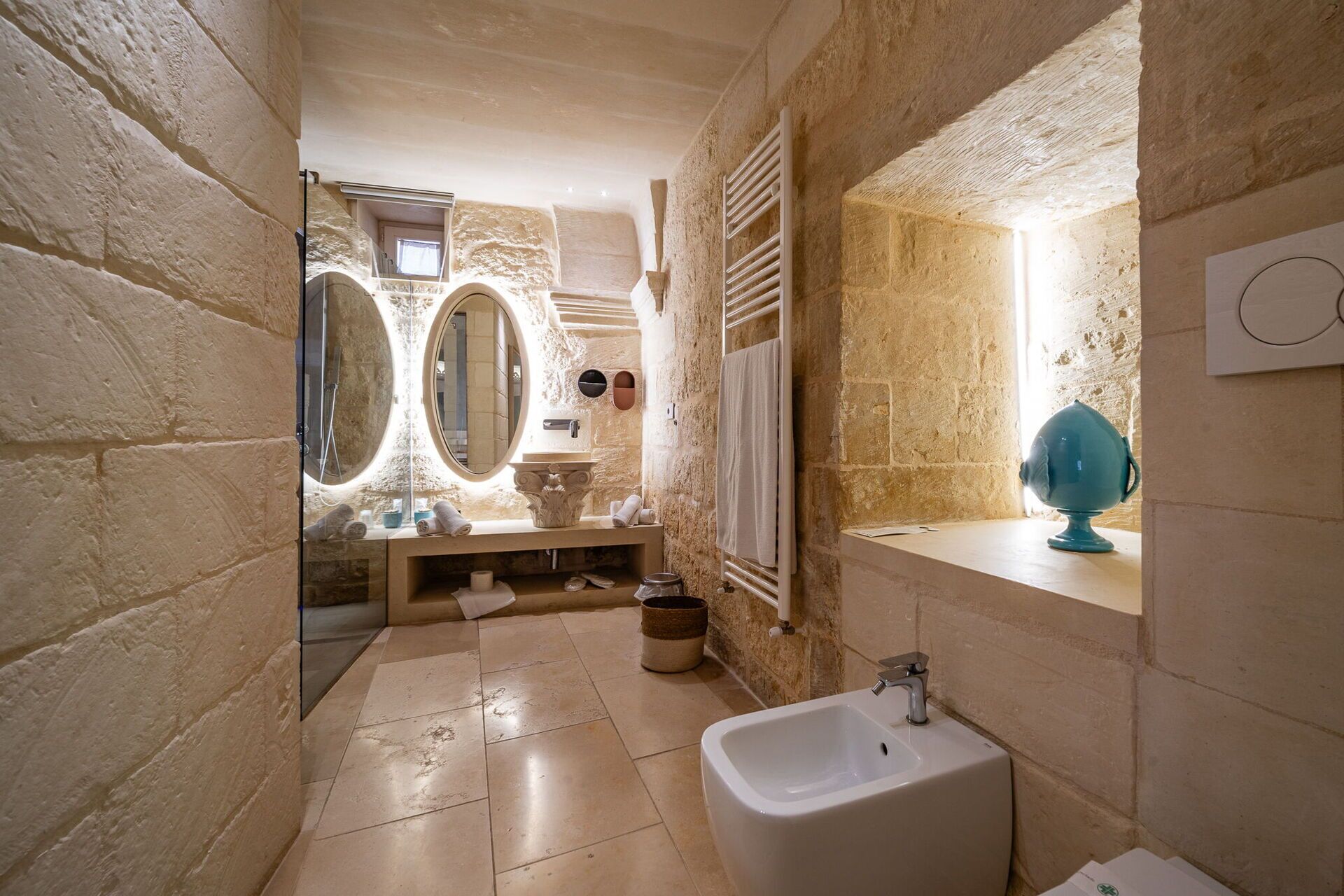 A narrow bathroom with stone walls, a sink, a bidet, and two large mirrors.