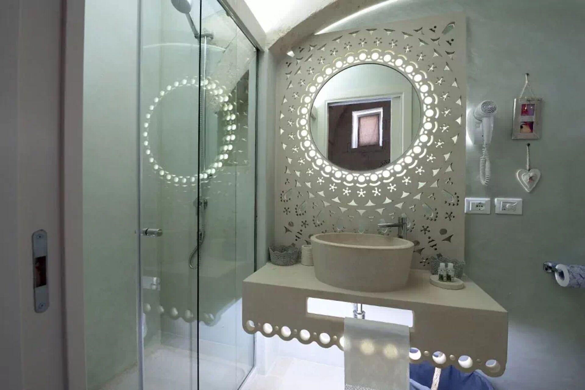 A bathroom with a glass shower enclosure, a vanity with a sink, a mirror with lights, and a wall-mounted telephone.