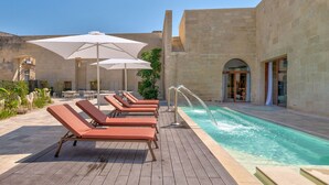 Ein Poolbereich mit Liegestühlen und Sonnenschirmen, ein klares Schwimmbecken und ein Steinhaus im Hintergrund.