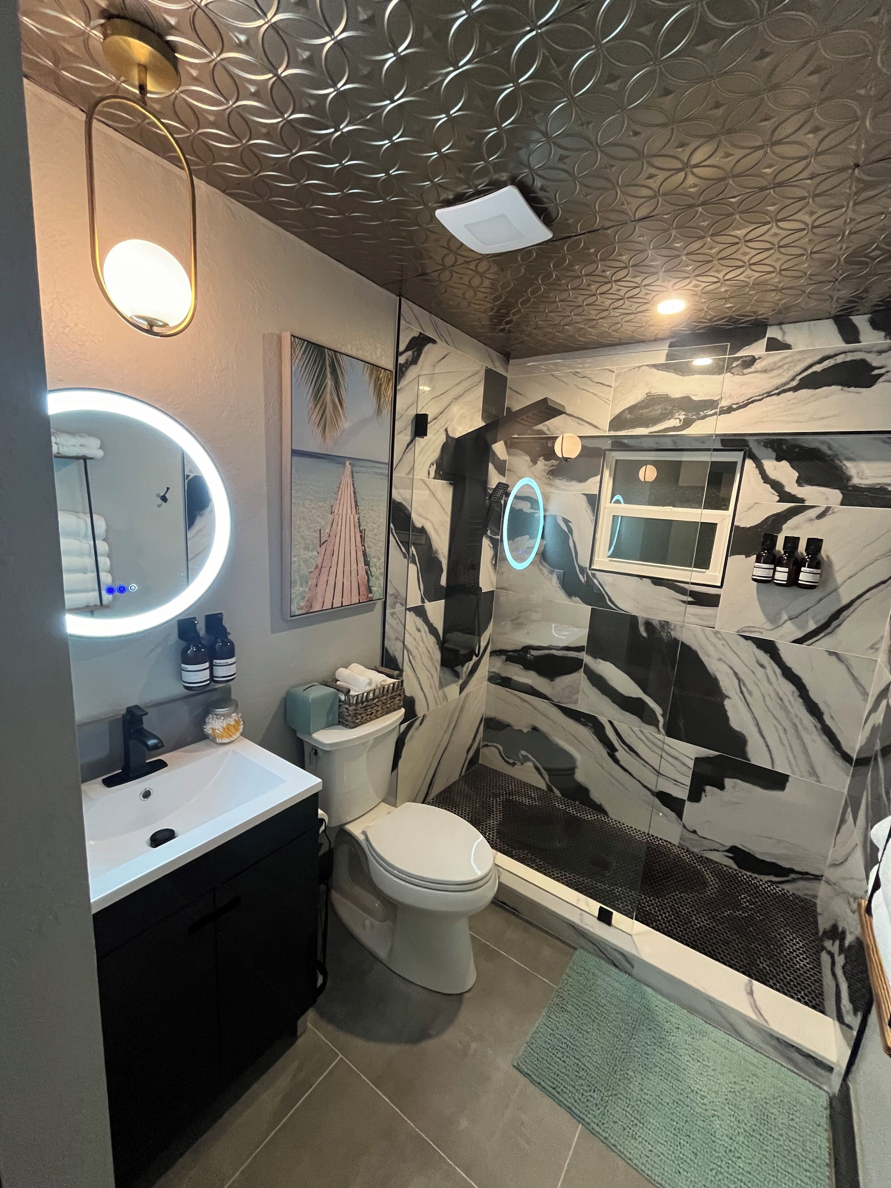 Une salle de bain au design moderne, dotée d’un grand miroir rond avec éclairage intégré, de toilettes, d’un lavabo et d’un espace douche avec un mur à motifs.