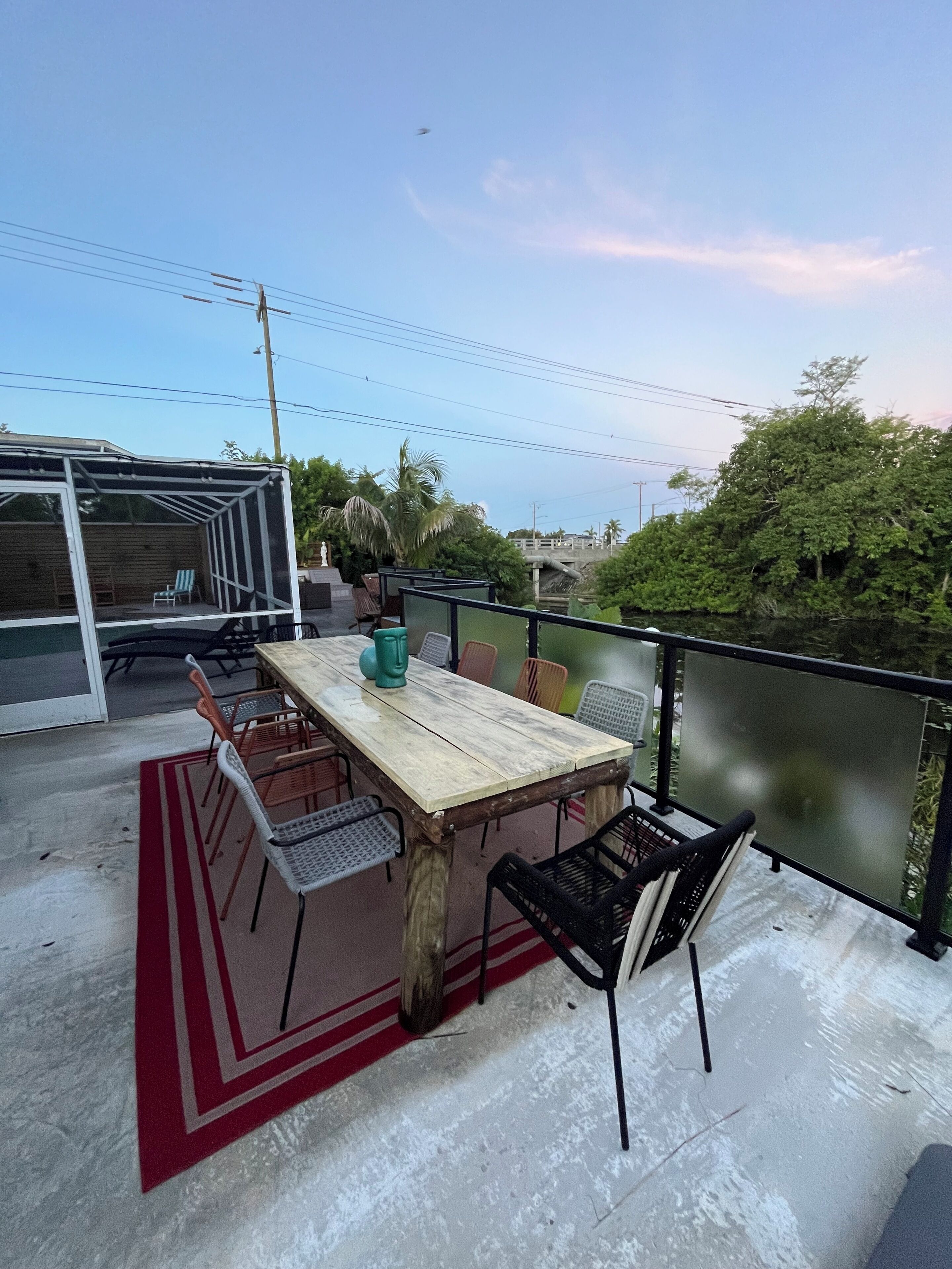 Une terrasse sur le toit avec une table et des chaises en bois, un tapis à rayures rouges et blanches, et une vue sur la verdure et un pont en arrière-plan.