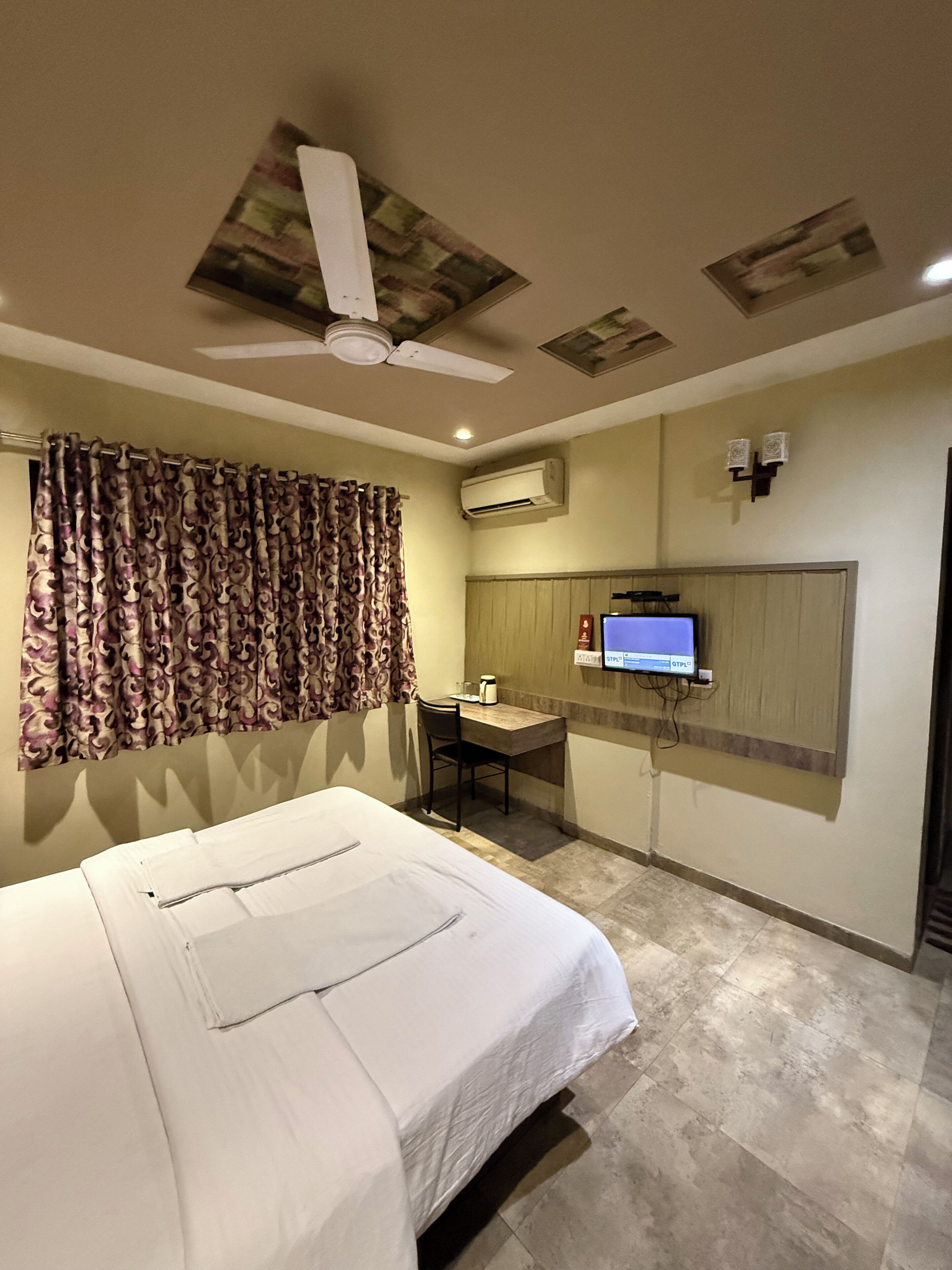 A hotel room with a bed, a desk, a chair, an air conditioning unit, a ceiling fan, and a television.
