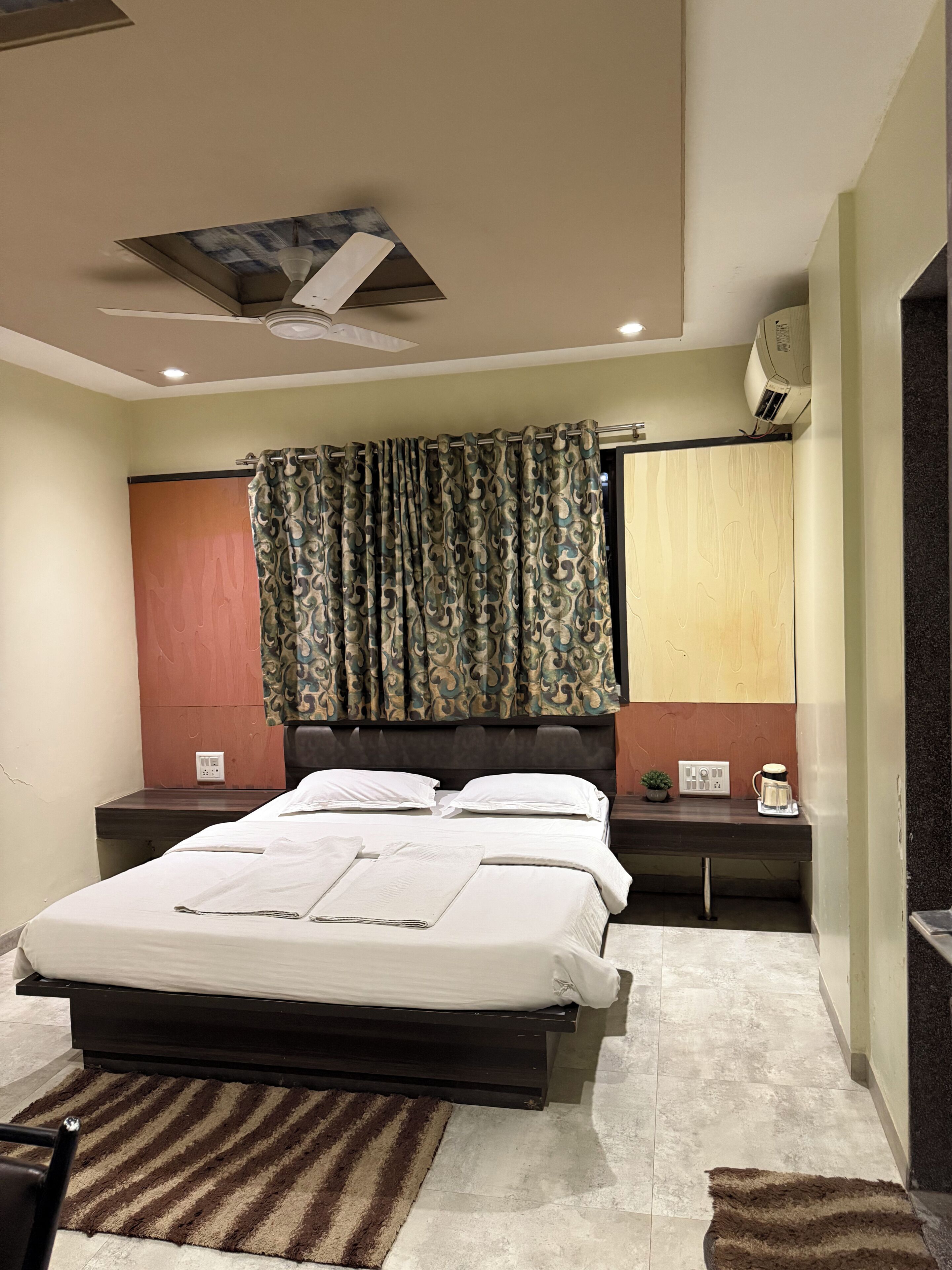 A bedroom with a bed, nightstands, a ceiling fan, a window with curtains, and a wall-mounted air conditioner.