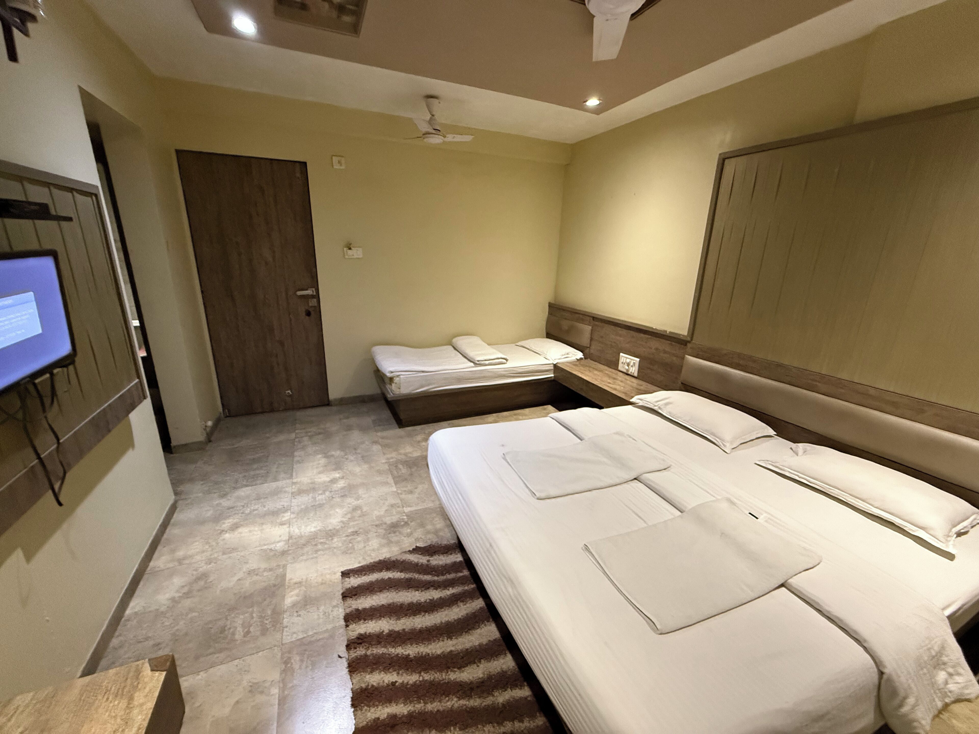 A hotel room with a large bed, a smaller bed, a desk, a chair, a TV, and a ceiling fan.