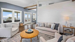 A spacious living room with a white sofa, a round wooden coffee table, and a ceiling fan. Large windows provide a view of the outdoors.