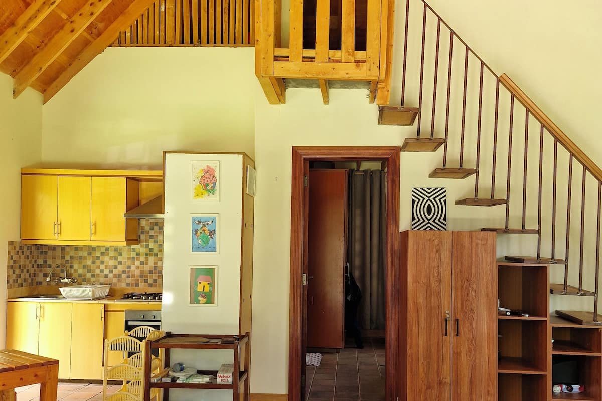 A spacious loft with a high ceiling, wooden beams, a kitchen with yellow cabinets, a dining area with a table and chairs, and a staircase with a wooden railing.