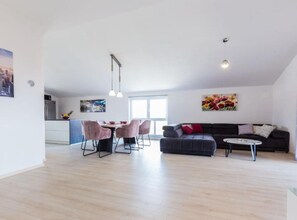 Un salón amplio con una mesa de comedor, sillas, un sofá y una mesa baja. Se ve una cocina al fondo con un refrigerador y un lavabo.