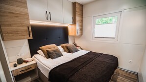 A bedroom with a bed, nightstands, a wooden headboard, a window, and a built-in wardrobe. - Lake Serenity Premium Mobile Homes With Thermal Riviera Tickets (Brežice)